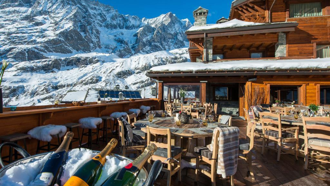 Un accogliente chalet di montagna a Cervinia vanta un patio soleggiato, arredato con tavoli e sedie. La neve ricopre i tetti e le cime circostanti, mentre le bottiglie di vino si raffreddano lì vicino. Il cielo azzurro brillante esalta questo scenario invernale.