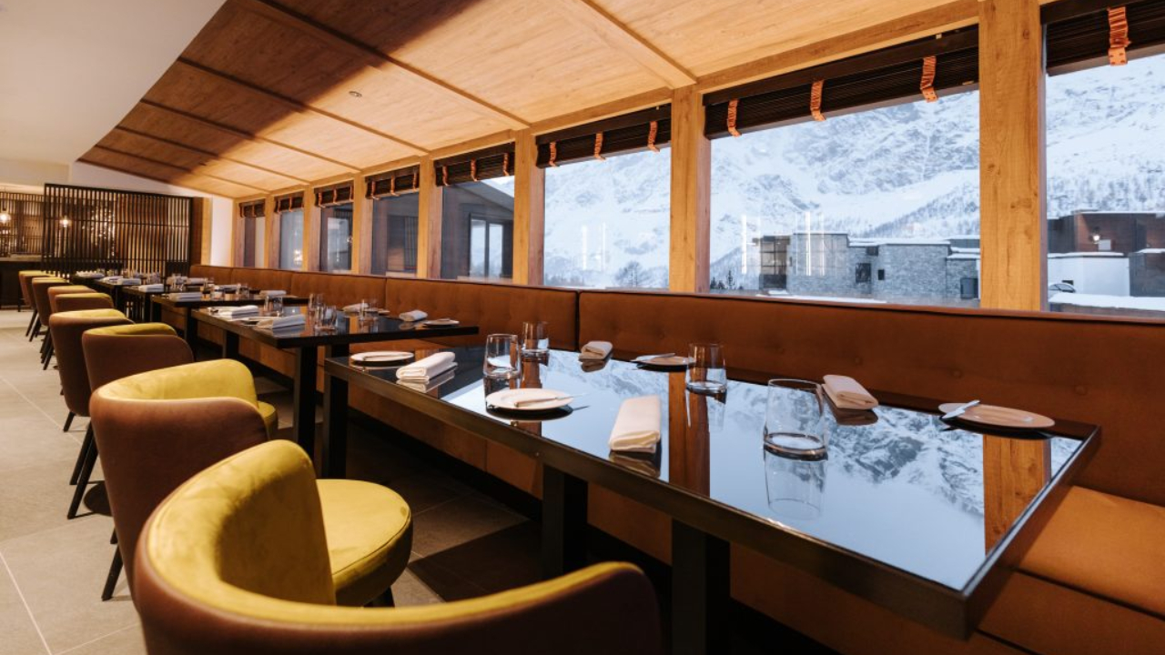 Un ristorante moderno con un caldo interno in legno presenta file di tavoli apparecchiati con bicchieri e tovaglioli. Sedie gialle e marroni fiancheggiano entrambi i lati, mentre grandi finestre incorniciano le splendide montagne innevate di Cervinia all'esterno.