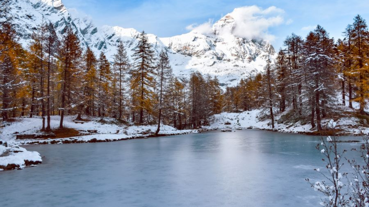 Un sereno lago ghiacciato vicino a Cervinia è circondato da alberi e montagne ricoperti di neve. Il cielo rimane limpido, con nuvole aggrappate a una cima innevata. Questo paesaggio mozzafiato trasuda un'atmosfera invernale pacifica.