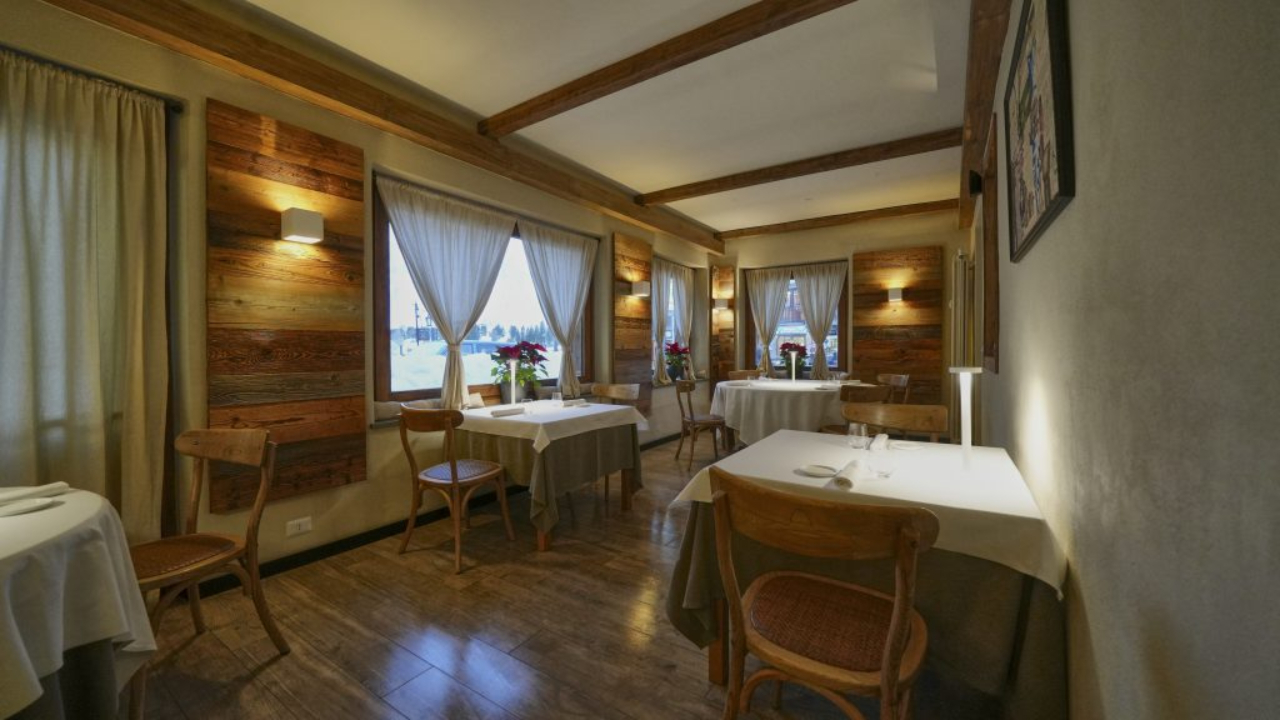 Un accogliente ristorante rustico a Cervinia presenta pareti in legno, travi a vista e tavoli con tovaglie bianche. Le sedie sono disposte ordinatamente attorno ai tavoli e la luce soffusa aggiunge calore. Le grandi finestre con tende lasciano entrare la luce naturale.