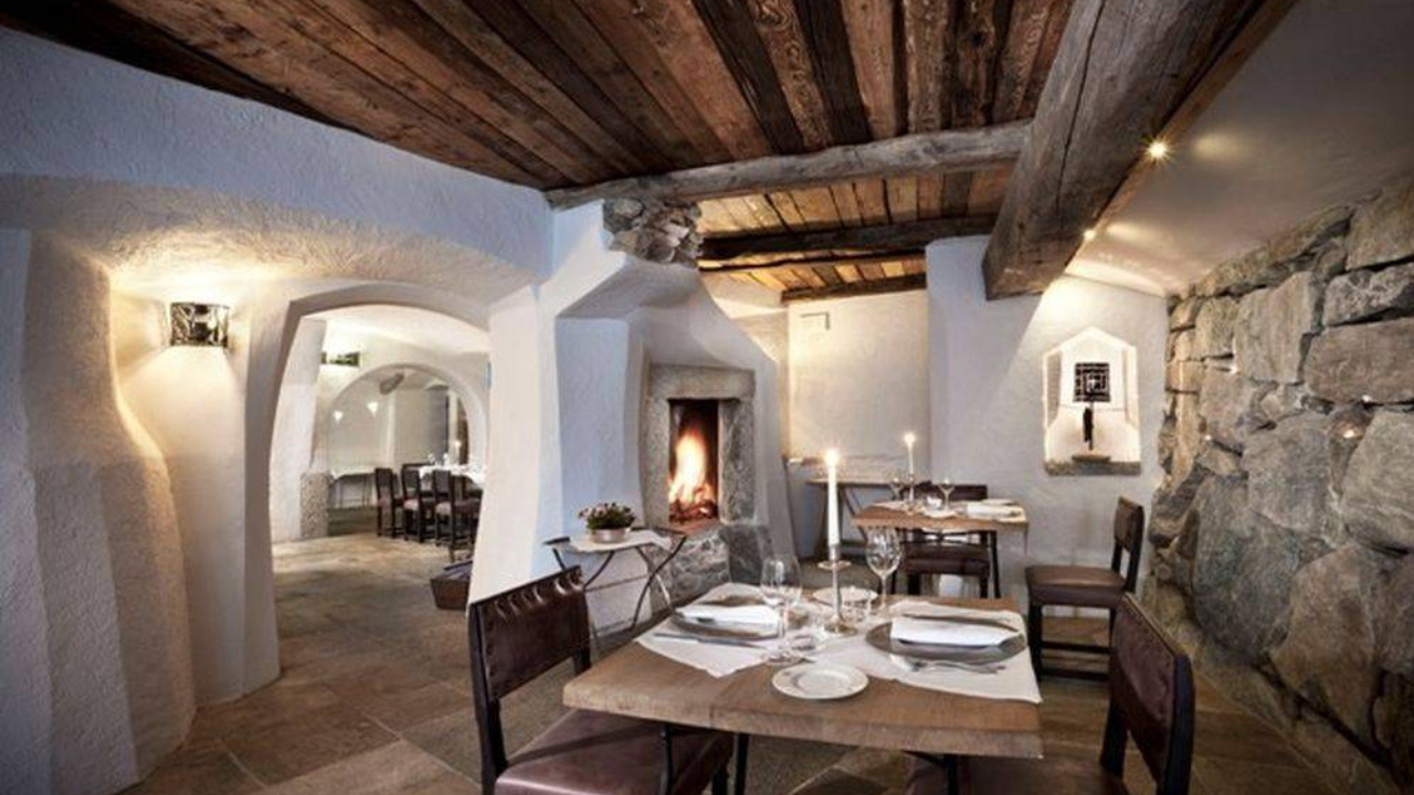 L'accogliente ristorante interno di Cervinia vanta pareti in pietra rustica, travi in legno sul soffitto e un camino acceso. Tavoli adornati con tovaglie bianche, stoviglie eleganti e candele ne esaltano il fascino. L'illuminazione ambientale diffonde un bagliore caldo e invitante in tutto lo spazio.