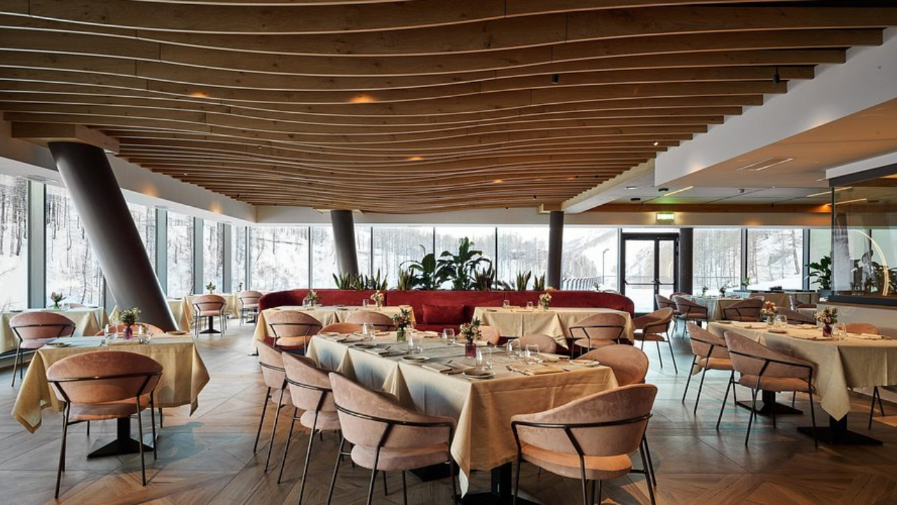 Un moderno ristorante interno a Cervinia vanta pannelli in legno sul soffitto e grandi finestre che incorniciano uno scenario innevato all'aperto. Sedie imbottite rosa, tavoli con tovaglie beige e piante decorative creano un'atmosfera accogliente in mezzo al fascino alpino.
