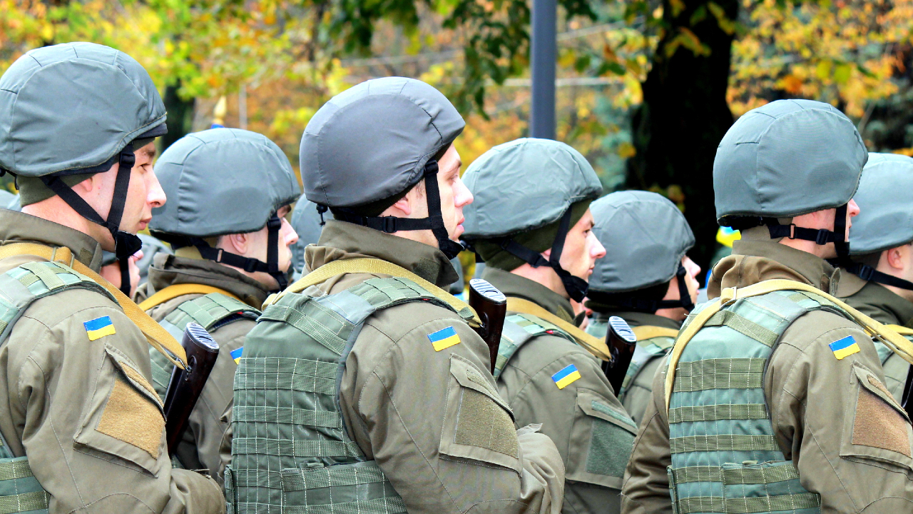 Un gruppo di soldati in uniforme è in formazione, con caschi e uniformi verdi ornati da toppe blu e gialle che mostrano con orgoglio la bandiera ucraina. Gli alberi si confondono sullo sfondo, alludendo al loro ambiente esterno.