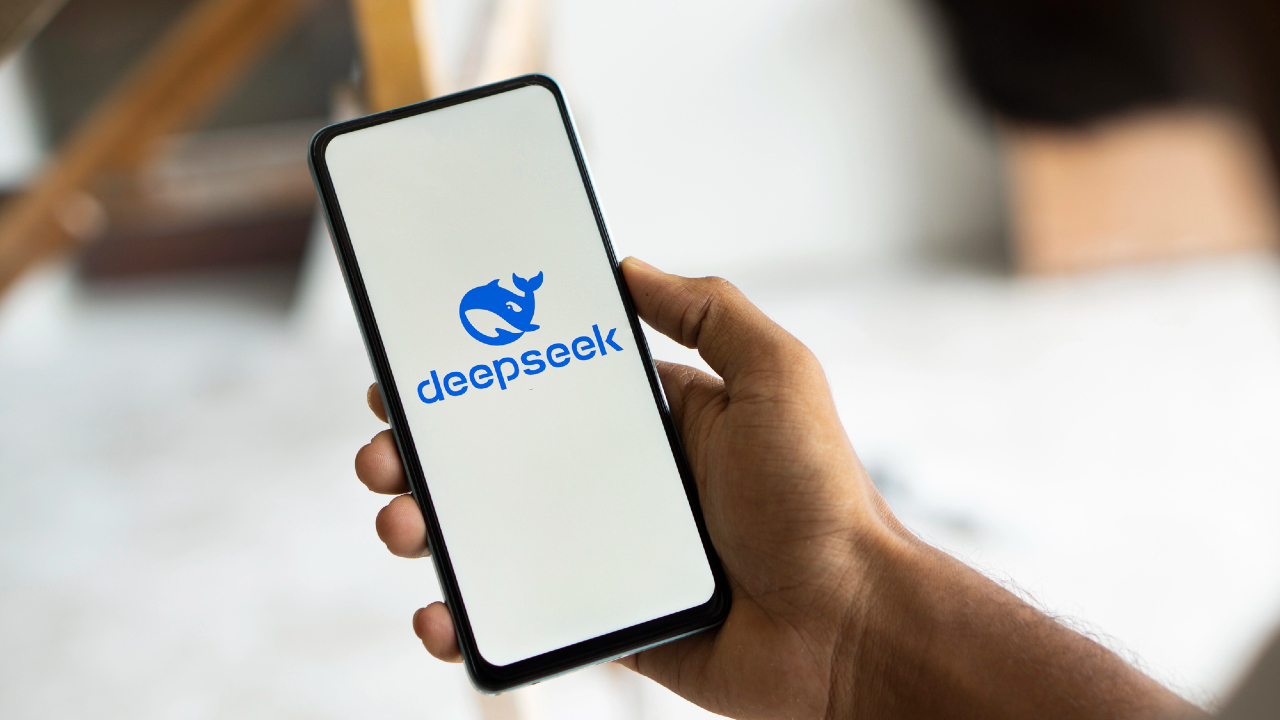 Una persona tiene in mano uno smartphone con un rinoceronte stilizzato blu con il testo "deepseek" su uno schermo bianco, a simboleggiare l'innovazione in azioni tech. Lo sfondo sfocato accentua lo schermo del telefono.