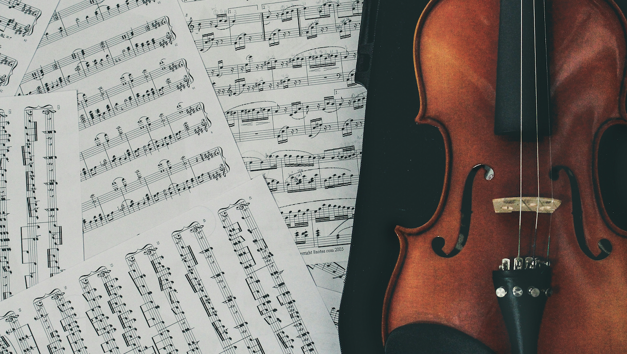Un violino di legno giace accanto a fogli sparsi di spartiti musicali. Le note musicali sono in inchiostro nero su carta bianca, creando un contrasto con i toni ricchi e caldi del legno del violino. La scena suggerisce un'ambientazione di musica classica.