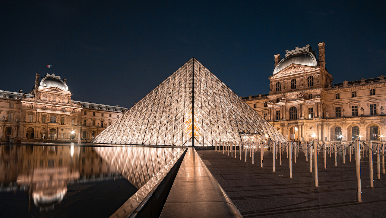 L'immagine mostra la Piramide del Louvre a Parigi, splendidamente illuminata di notte. Si trova di fronte allo storico Palazzo del Louvre, con le sue luci che si riflettono sull'acqua che circonda la piramide. Il cielo è scuro, aggiungendo contrasto.