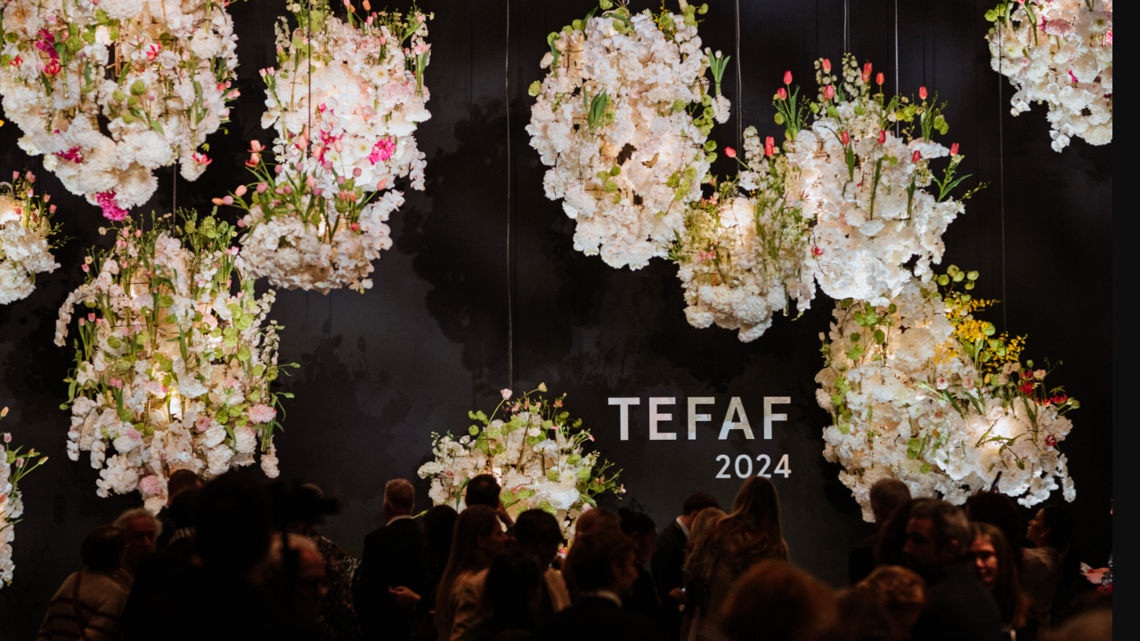 Composizioni floreali sospese con fiori bianchi e accenni di rosa e giallo sono esposte su uno sfondo scuro. Sul muro è scritto "TEFAF 2024". Una folla di persone è radunata sotto.