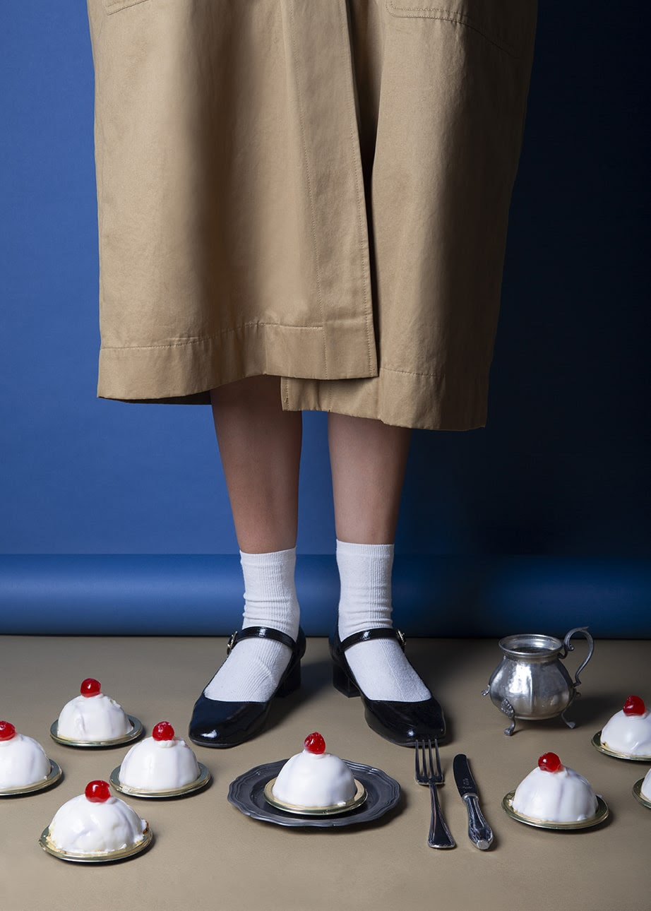 Una persona con gonna beige e scarpe nere con calzini bianchi, incarnando l'estetica di Manfrone, è in piedi in bilico davanti a uno sfondo blu. Diverse piccole torte guarnite con ciliegie sono disposte ad arte sul pavimento, accompagnate da una forchetta, un coltello e una piccola teiera metallica.