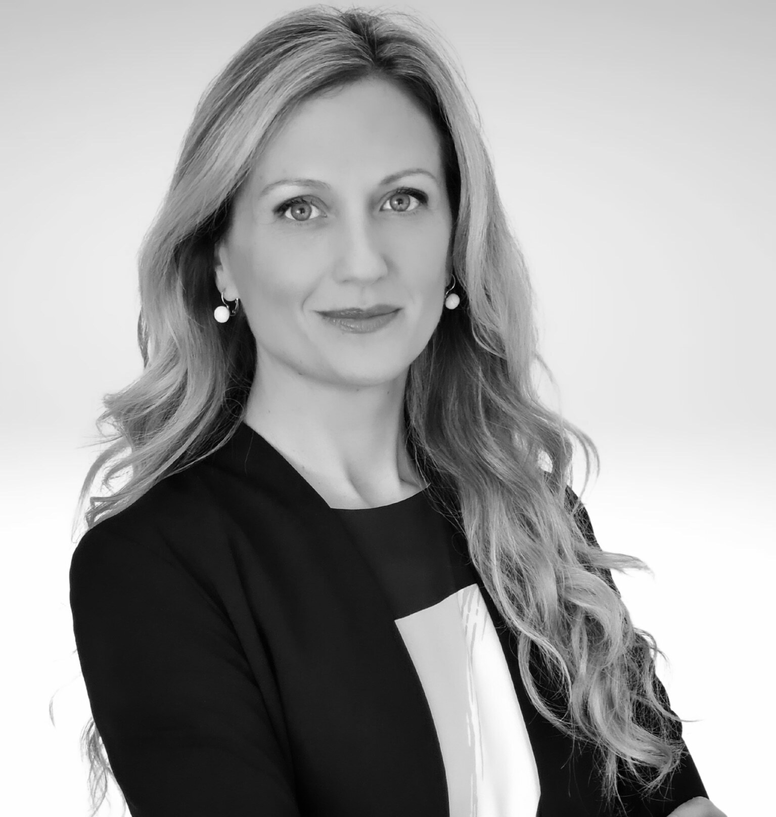 Una donna con lunghi capelli ondulati, che indossa un blazer scuro e un top chiaro, è in piedi su uno sfondo semplice. È rivolta in avanti e sorride dolcemente. L'immagine è in bianco e nero.