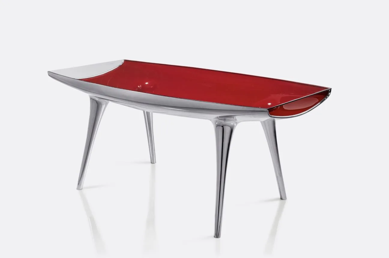 Un tavolo moderno con un'elegante struttura in argento metallizzato e quattro gambe angolari. Il piano è rosso lucido con bordi curvi, simili a quelli di una barca, che gli conferiscono un aspetto artistico e futuristico.