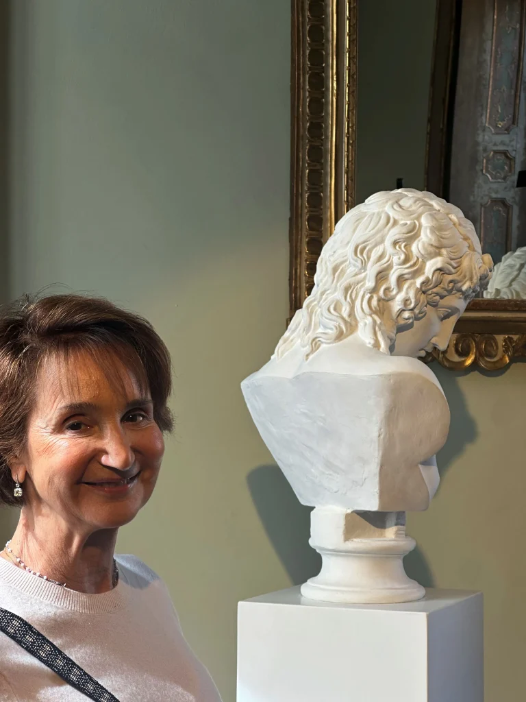 Una donna con i capelli castani corti è in piedi e sorridente accanto a un busto di marmo bianco su un piedistallo, il cui riflesso è catturato in uno specchio incorniciato, evocando lo stile raffinato di Bassetti spesso visto agli eventi Thinking Italian di Christie's.