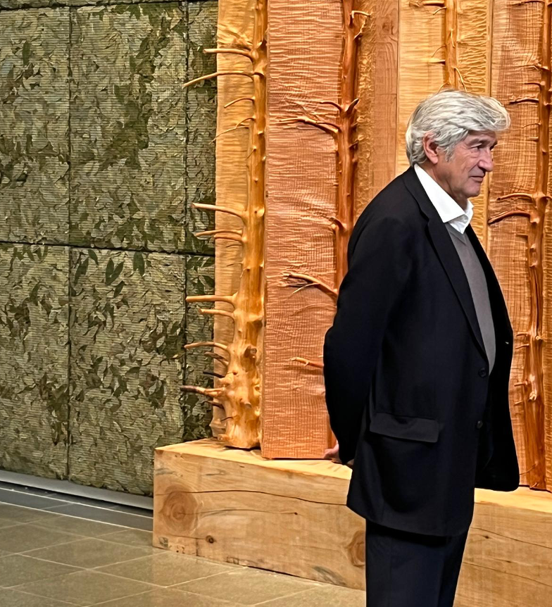 Un uomo con i capelli grigi, che indossa un abito nero e una camicia bianca, è in piedi in un interno, davanti a una grande scultura in legno che ricorda tronchi d'albero, posta contro una parete verde testurizzata.