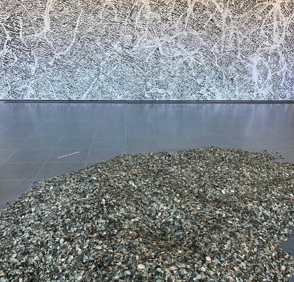 Un grande mucchio di gusci di ostriche è sparso su un pavimento di piastrelle grigie di fronte a una parete ricoperta di intricati disegni astratti in bianco e nero che ricordano rami o radici di alberi.