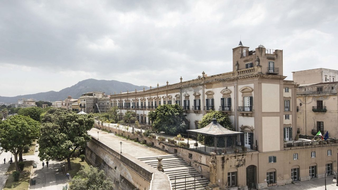 Un grande edificio storico con colonne e dettagli decorati sorge accanto a una strada alberata e a giardini a Palermo, con montagne visibili sullo sfondo sotto un cielo nuvoloso.