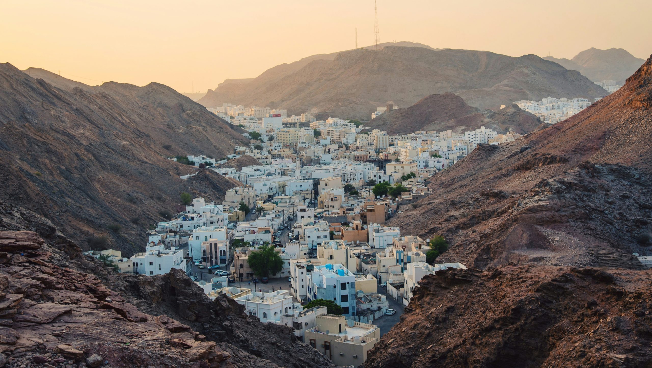 Una città con edifici bianchi incastonata tra montagne rocciose e marroni, sotto un cielo arancione e nebbioso al tramonto: uno scenario ideale per un viaggio in Oman. Le montagne incorniciano le case a grappolo, con tocchi di verde tra gli edifici.