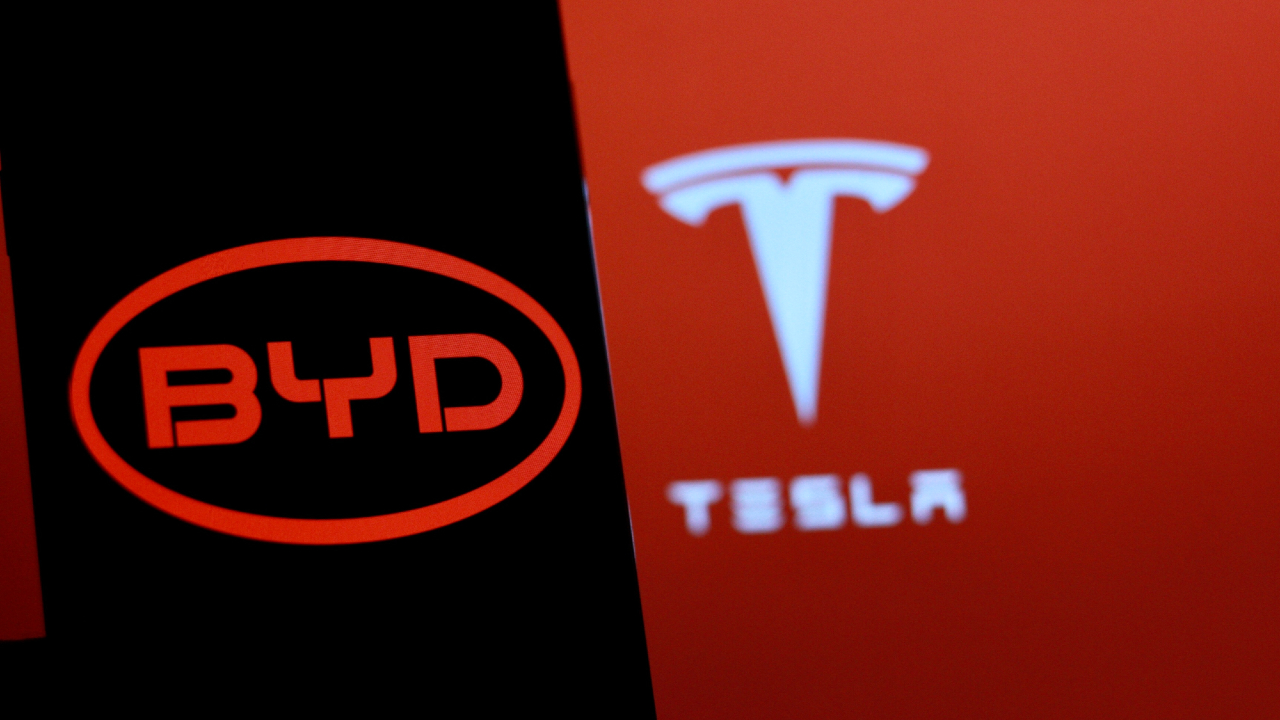 Un'immagine sorprendente in rosso e nero presenta due loghi iconici: sulla sinistra, BYD si erge audacemente in un ovale rosso su uno sfondo nero; sulla destra, l'elegante logo a "T" e il nome di Tesla sono elegantemente visualizzati in bianco su uno sfondo rosso brillante.