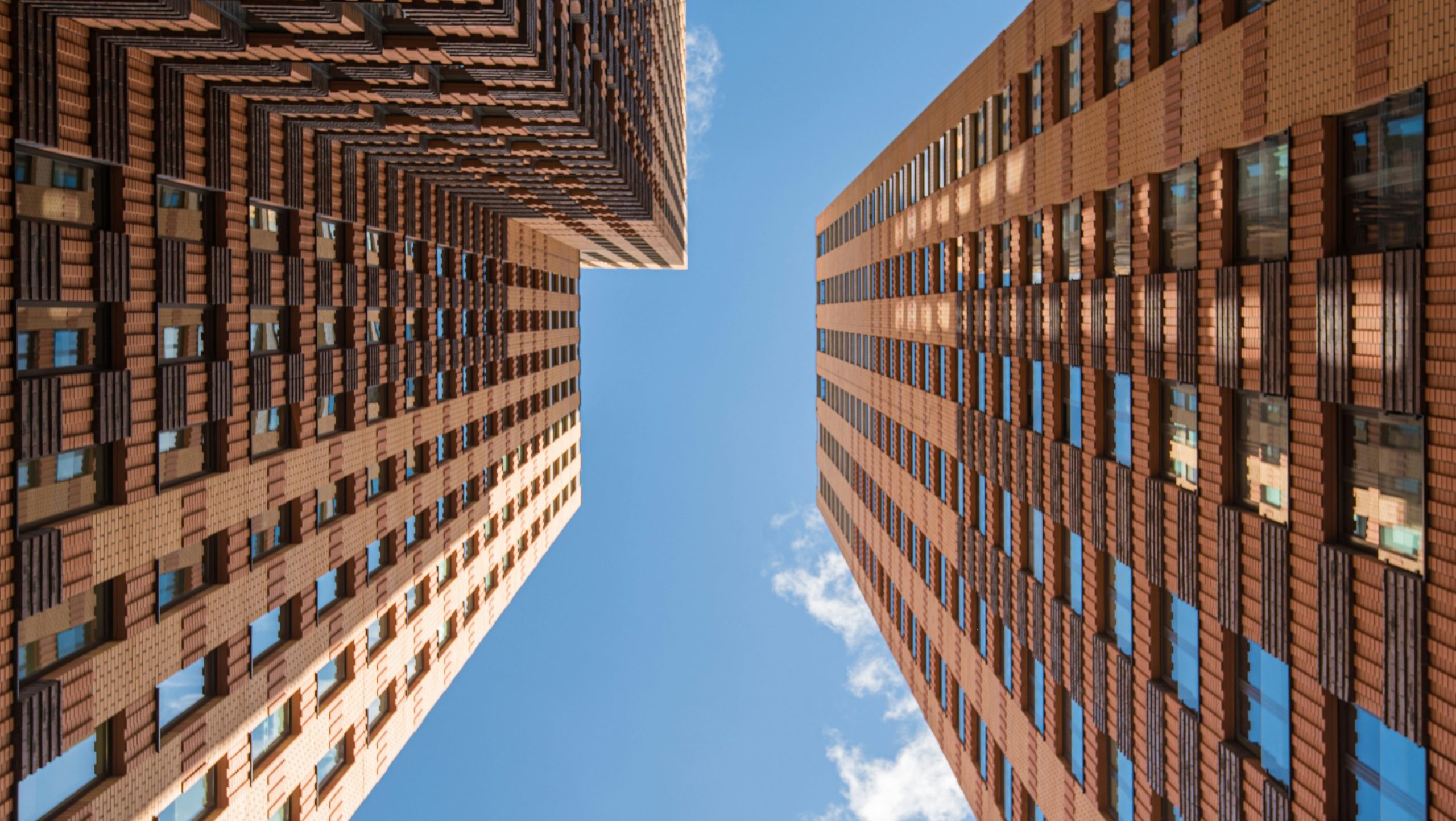 Due alti e moderni edifici in mattoni si ergono su entrambi i lati, visti dal basso, con il cielo azzurro e qualche nuvola visibile tra di essi. I motivi ripetuti delle finestre e i disegni geometrici conferiscono un aspetto distintivo, che ricorda l'architettura di TFM Polizza.