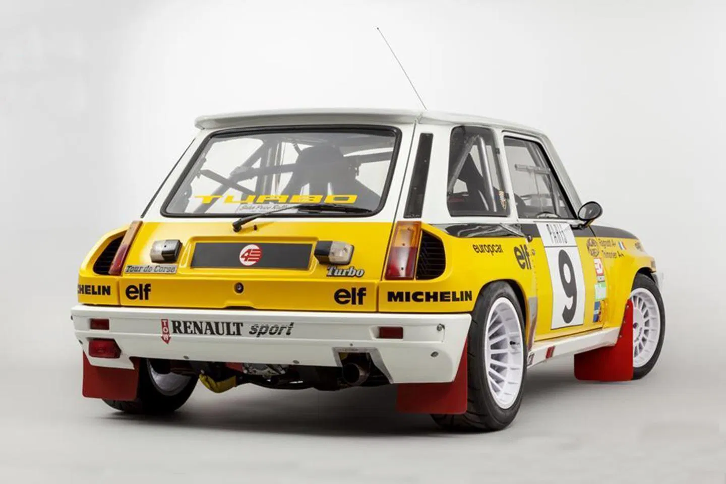 Una classica auto da rally Renault 5 Turbo gialla e bianca con adesivi da corsa, numero 9 sulla portiera e spoiler posteriore, vista da dietro su uno sfondo bianco.