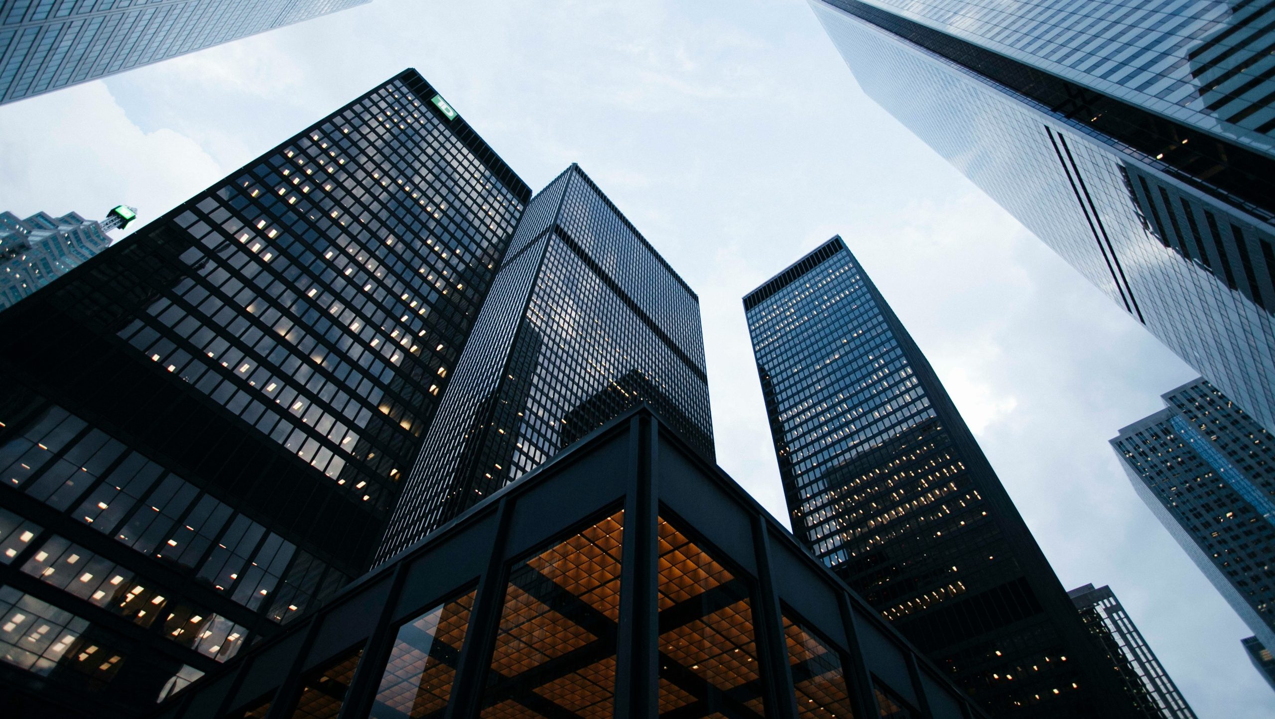 Vista di alti e moderni grattacieli da un'angolazione bassa, che crea una spettacolare prospettiva verso l'alto contro un cielo nuvoloso. Gli edifici sono caratterizzati da vetro e acciaio riflettenti, contribuendo a creare un elegante paesaggio urbano.