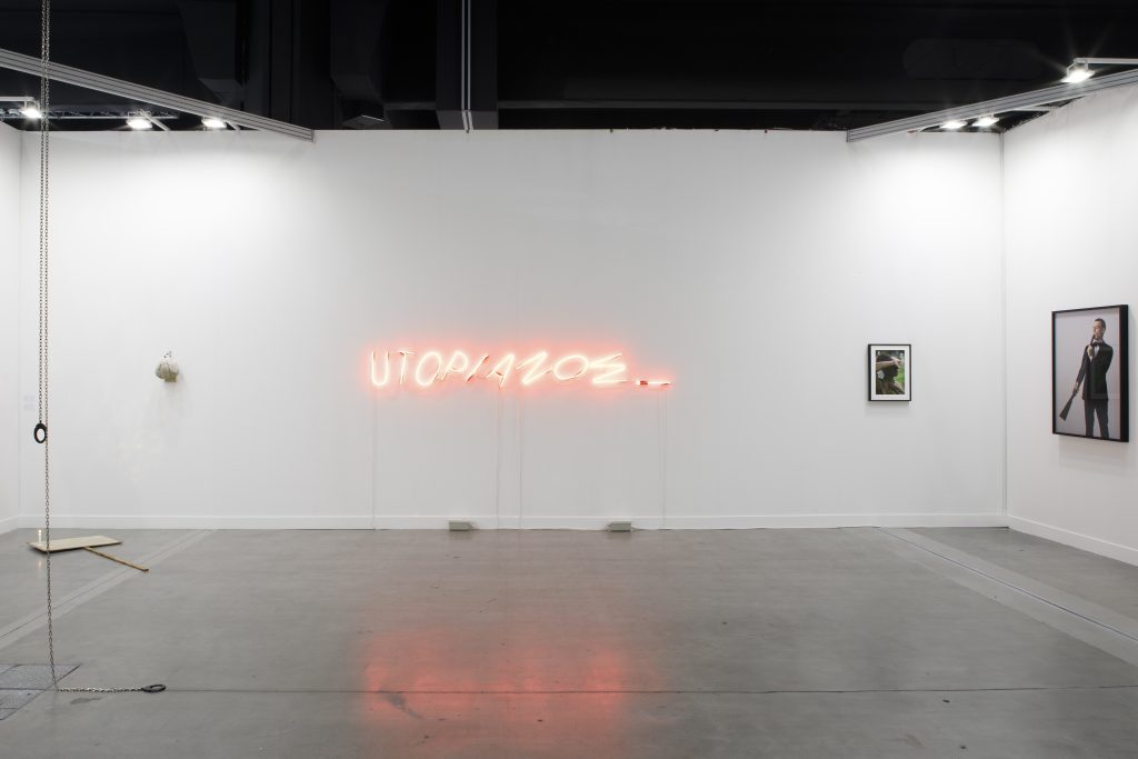 In questa galleria minimalista e industriale, un'insegna al neon irradia "UTOPIA 2023". Sulla destra, due opere d'arte incorniciate catturano i visitatori, mentre oggetti eclettici e una corda oscillante amuletano sulla sinistra. Questo spazio intrigante prepara il terreno per la prossima mostra miart 2025 presso Gallerie Utopia.
