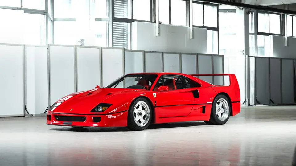 Una Ferrari F40 rossa brillante è parcheggiata all'interno su un pavimento lucido con ampie finestre e pareti bianche sullo sfondo. L'auto ha un design elegante e basso e un alettone posteriore prominente.