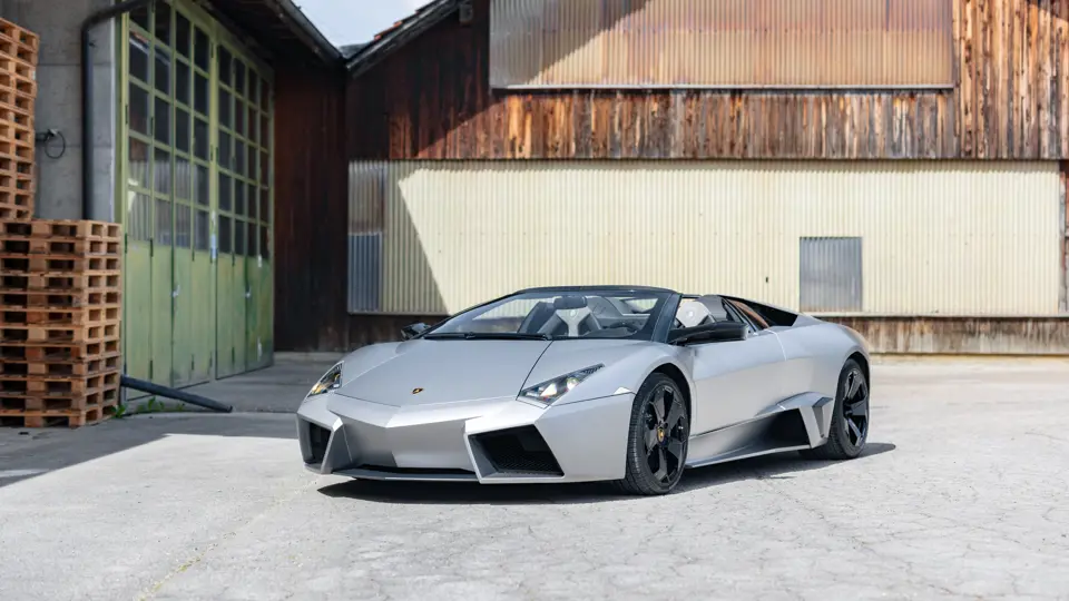 Una Lamborghini Reventón Roadster argentata è parcheggiata all'esterno di un edificio rustico con pareti in legno e metallo, vicino a pallet di legno accatastati.