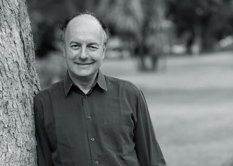 A man with short hair and a receding hairline, wearing a button-up shirt, stands next to a tree and smiles at the camera in a park setting. The photo is in black and white.