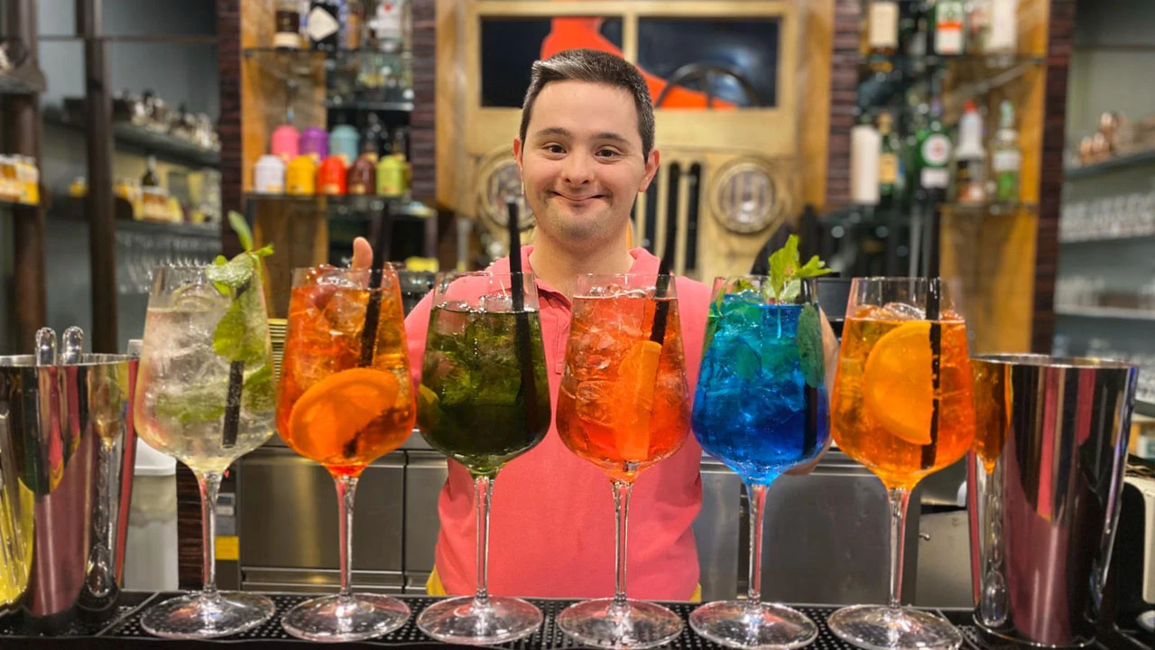 Un barista sorridente con una camicia rosa è in piedi dietro una fila di cocktail colorati guarniti con frutta e menta in un bar ben fornito, pronto a mostrare agli ospiti come gustare i loro drink como un verdadero experto.