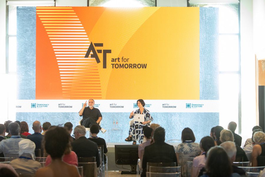 Un uomo e una donna parlano sul palco davanti al pubblico durante un evento di Art for Tomorrow, incorniciati da un grande cartello arancione e giallo con la scritta "Art for Tomorrow". La sala illuminata è piena di partecipanti rivolti verso i relatori.