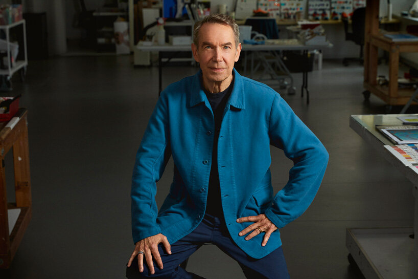 Una persona che indossa una giacca blu e pantaloni scuri posa con sicurezza in uno studio d'arte, circondata da tavoli da lavoro e materiali creativi, incarnando lo spirito dell'arte del futuro.