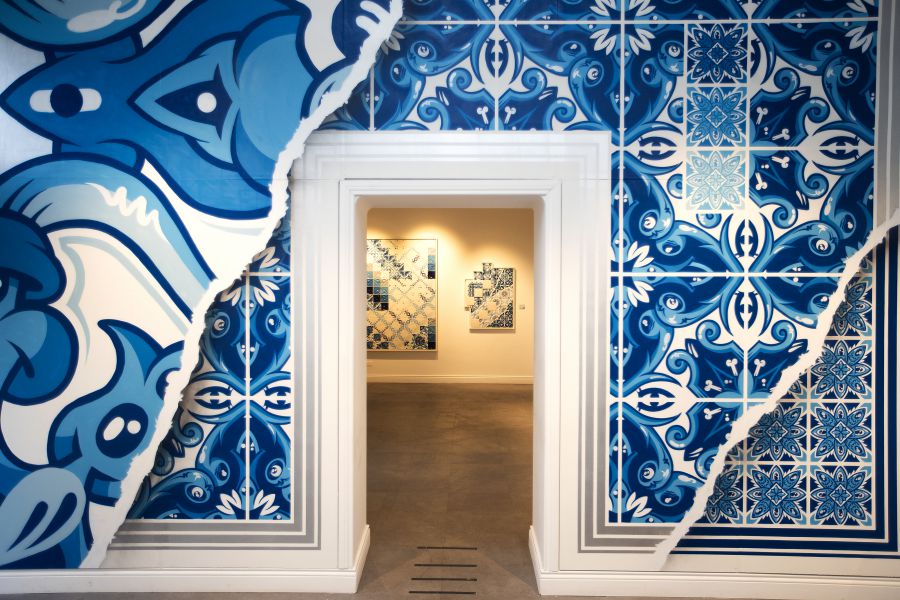 Un portale incastonato in una parete decorata con piastrelle blu e bianche ispirate al deodato, parzialmente strappate per rivelare l'interno di una moderna galleria con opere d'arte con motivi simili esposte su pareti beige.