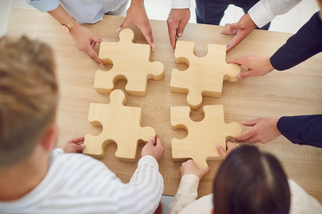 Sei persone sedute attorno a un tavolo tengono ciascuna un grande pezzo di puzzle in legno e lavorano insieme per incastrare i quattro pezzi al loro posto, a simboleggiare il lavoro di squadra e la collaborazione.
