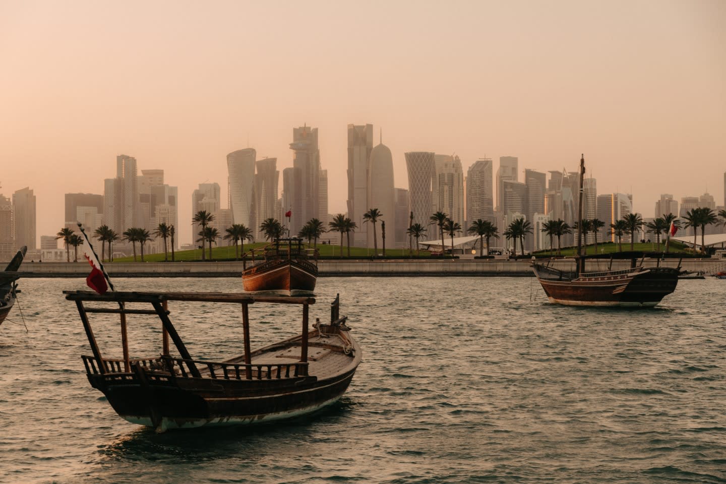 Le tradizionali barche di legno galleggiano sulle acque calme, con una fila di palme e il moderno skyline della città di Doha, sede di Art Basel, sullo sfondo, sotto un cielo nebbioso al tramonto.