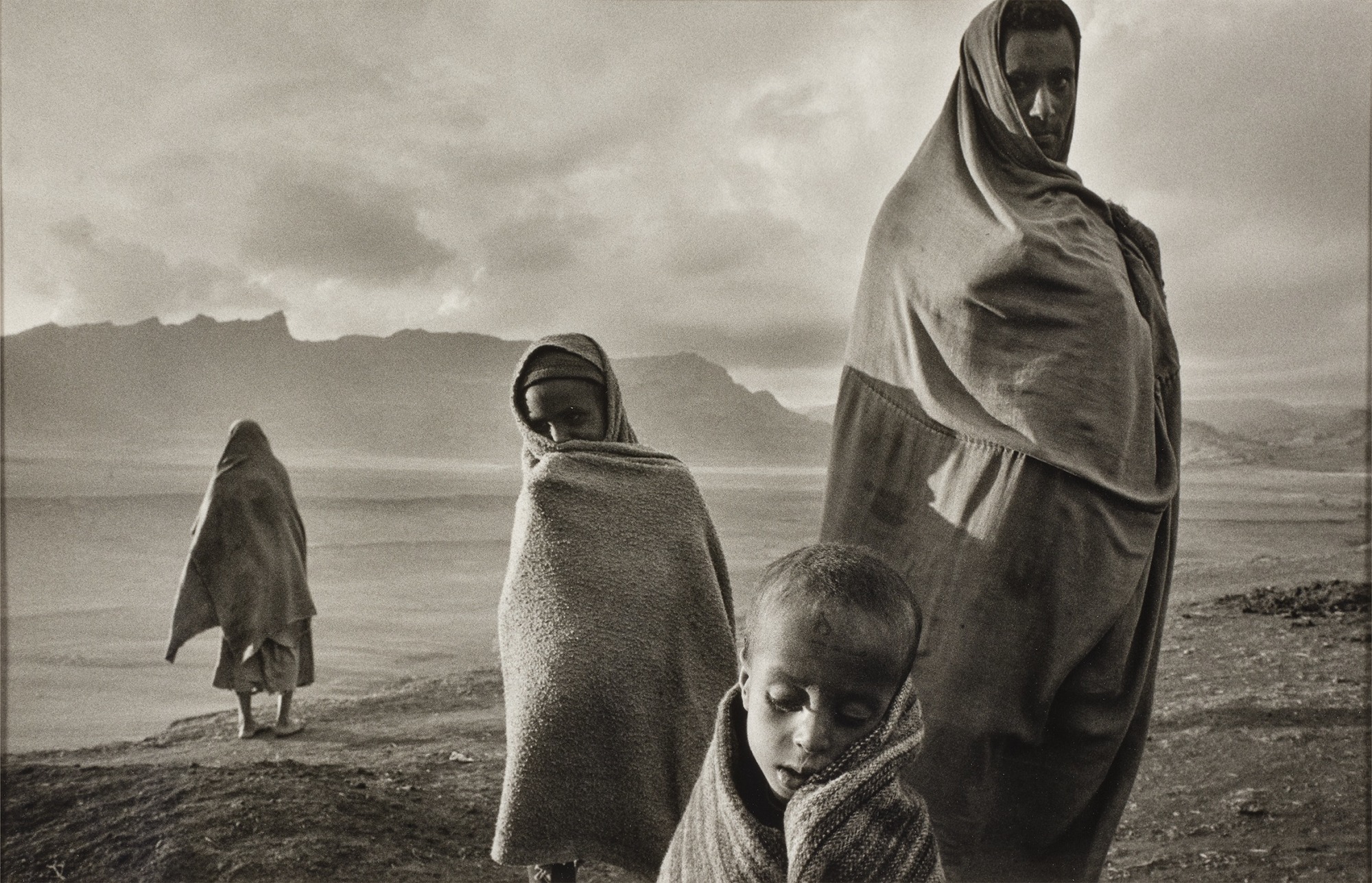 Quattro persone, tra cui bambini, sono all'aperto avvolte in coperte, con un paesaggio desolato e montagne sullo sfondo. L'immagine è in bianco e nero, evocando un'atmosfera cupa e riflessiva.