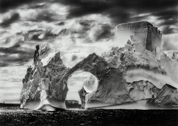 Una spettacolare foto in bianco e nero di un iceberg frastagliato con un'apertura ad arco, che galleggia in acque scure sotto un cielo nuvoloso e granuloso. Il ghiaccio imponente mostra dettagli intricati e forti contrasti di luce e ombra.