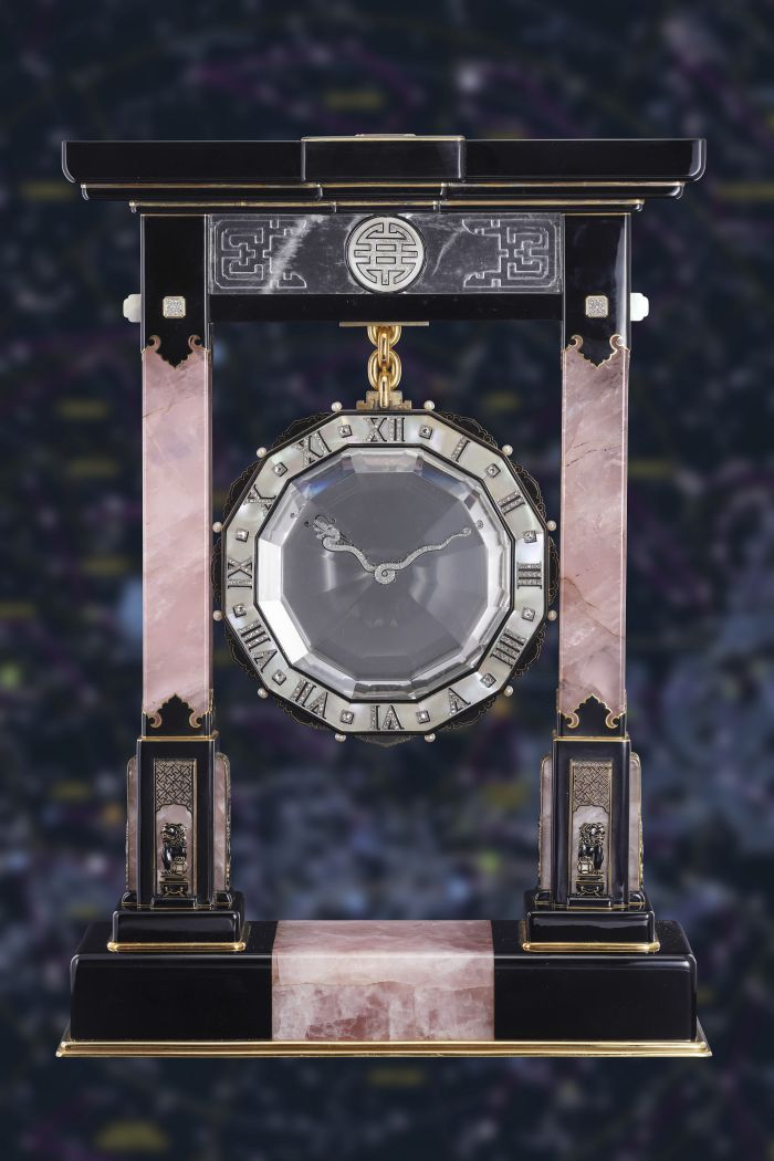 Un orologio decorativo con quadrante esagonale, numeri romani e lancette decorate, un esempio di orologi lusso nuove tendenze, sospeso tramite una catena d'oro tra due colonne di marmo rosa e nero, su uno sfondo scuro e sfocato.