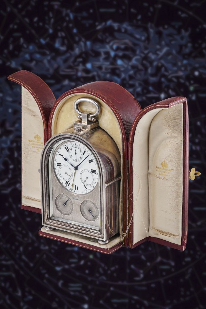 Un orologio da viaggio vintage in argento con due quadranti, che riflette gli orologi lusso nuove tendenze, è esposto in una custodia aperta in pelle rossa foderata con tessuto color crema, su uno sfondo scuro e stellato.