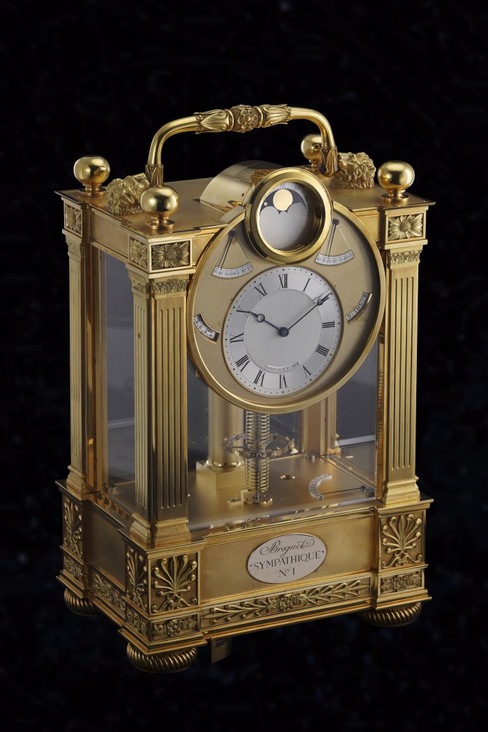 Un lussuoso orologio da carrozza in oro con colonne ornate, motivi floreali e lati in vetro trasparente mette in mostra gli orologi di lusso "Nuove Tendenze". Il quadrante dettagliato reca l'etichetta "Breguet Sympathique N. 1", su uno straordinario sfondo nero.