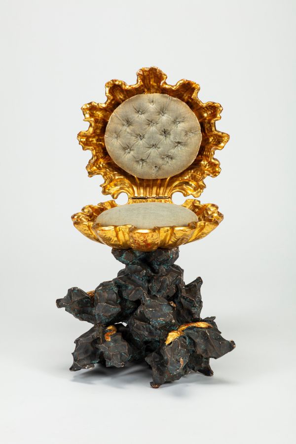Una sedia decorata che ricorda una conchiglia dorata con un rivestimento capitonné chiaro è appoggiata su una base scura e frastagliata, simile a una roccia, su uno sfondo chiaro e semplice: un design che ricorda l'estetica di Stefano Gabbana nell'ultima collezione Asta.