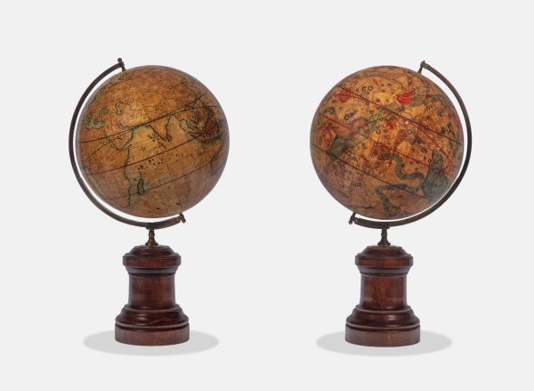 Due antichi mappamondi con mappe vintage dettagliate, tra cui una mappa celeste, sono esposti uno accanto all'altro su supporti di legno scuro su uno sfondo bianco semplice, evocando l'eleganza senza tempo di una collezione Asta di Stefano Gabbana.