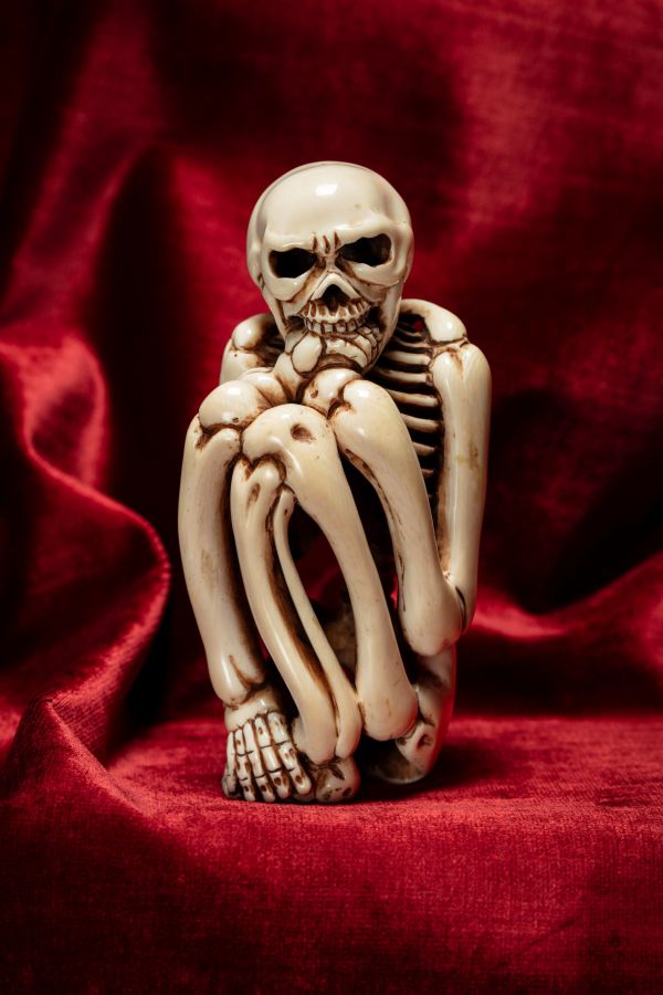 Una piccola statuina di scheletro pallido è seduta con le ginocchia raccolte al petto su uno sfondo di velluto rosso intenso, che ricorda lo stile drammatico delle collezioni Asta di Stefano Gabbana.