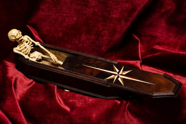 Una piccola bara di legno con intarsi a forma di rosa dei venti, parzialmente aperta per rivelare uno scheletro umano al suo interno, evoca il mistero dell'asta di Stefano Gabbana, il tutto adagiato su un ricco tessuto di velluto rosso.