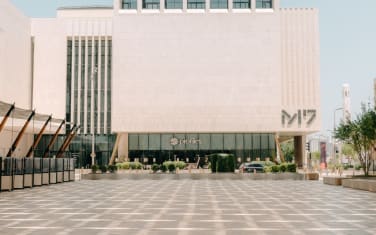 Un edificio moderno con grandi muri in pietra beige e alte finestre, contrassegnato con la scritta "M7", sorge dietro una spaziosa piazza decorata con alberi e alcune auto parcheggiate, evocando l'energia creativa di Art Basel Doha.