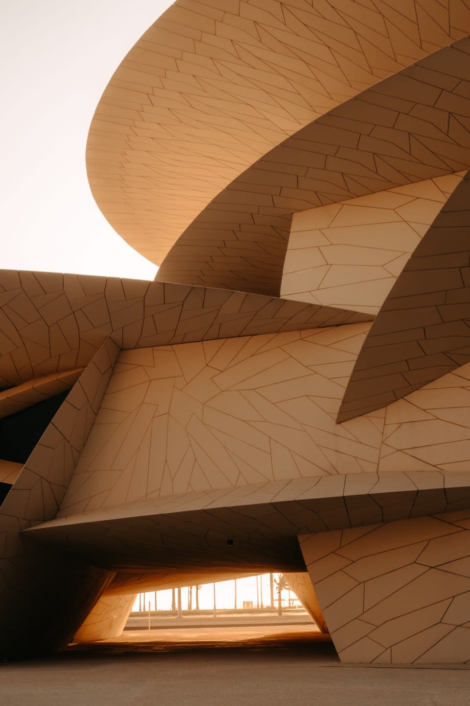 Moderna struttura architettonica con pannelli geometrici curvi e sovrapposti nei toni del beige, che ricordano dischi interconnessi. Ricordando le installazioni di Art Basel Doha, la luce del sole filtra attraverso la base, proiettando un bagliore caldo e ombre spettacolari.