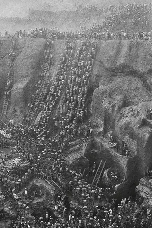 Centinaia di persone con indosso un cappello scalano ripide e accidentate pareti di terra, salendo su scale e camminando in file serrate, come se stessero lavorando intensamente in un immenso sito di scavi a cielo aperto.