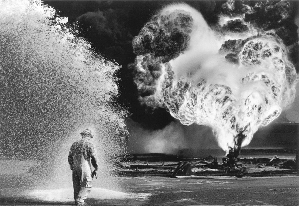 Una persona con equipaggiamento protettivo viene spruzzata di schiuma nei pressi di un grande incendio in eruzione e di un fumo denso, probabilmente in un giacimento petrolifero o in un sito industriale, riecheggiando le scene drammatiche catturate da Salgado, con caotiche colonne di fumo che si alzano nel cielo.