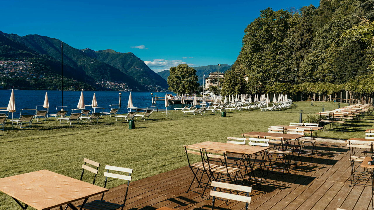 Tavoli e sedie in legno sono disposti su un prato erboso vicino al Lago di Como, con file di lettini e ombrelloni bianchi. Gli alberi costeggiano l'area e le montagne si stagliano sullo sfondo sotto un cielo azzurro e terso.
