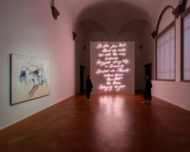 Una galleria d'arte scarsamente illuminata presenta una grande insegna al neon luminosa con scritte in corsivo sulla parete di fondo e un dipinto astratto sulla parete di sinistra. Due persone, in disparte, osservano le opere d'arte nell'ampia sala.