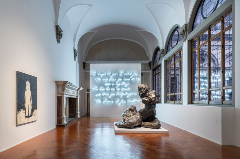 Una galleria d'arte contemporanea con alti soffitti a volta presenta un dipinto in bianco e nero di una ragazza, una grande scultura astratta e un testo corsivo al neon che illumina una parete bianca. Alte finestre fiancheggiano un lato dello spazio.