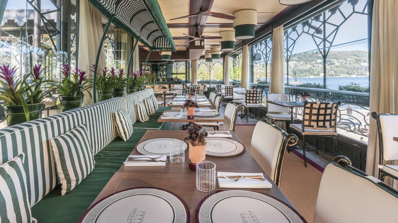 Un ristorante luminoso ed elegante, con cuscini a righe verdi e bianche, tavoli apparecchiati, piante in vaso e ampie finestre che si affacciano sulle splendide acque e sulle colline del Lago di Como nelle giornate di sole.